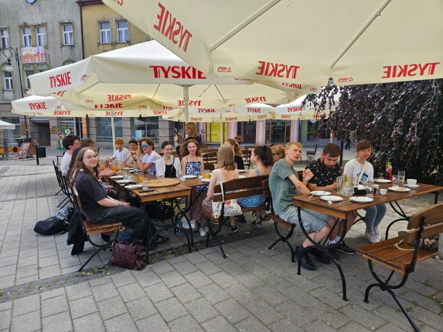 Grupa młodych osób siedzi przy długim stole w ogródku restauracyjnym na świeżym powietrzu. W tle widać kamienice i deptak w miejskim rynku.